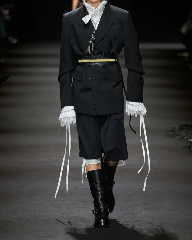 Модель Ann Demeulemeester в черном двубортном жакете, шортах и черных сапогах с ремнем и заклепками