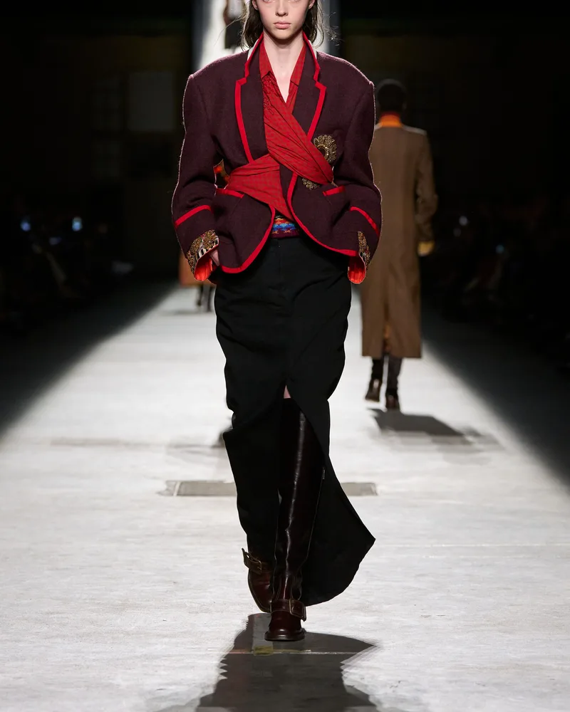Модель Dries Van Noten в бордовом жакете с красной отделкой, черной юбке и высоких сапогах с пряжкой